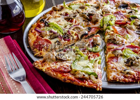 Close up of fresh baked thin crust supreme pizza surrounded by containers of red wine vinegar and olive oil with striped napkin and fork
