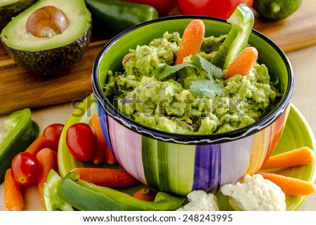 Fresh chunky guacamole in colorful bowl sitting on bright green plate garnished with raw carrots and green peppers and cilantro surrounded by raw vegetables