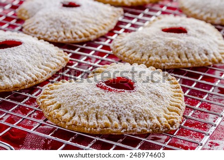 Heart shaped cherry hand pies dusted with powdered sugar sitting on wire cooling rack on top of red and white heart kitchen towel