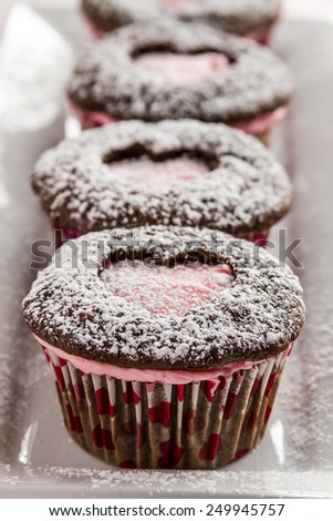 Close up of line of chocolate cupcakes with heart shaped cutout filled with pink frosting sitting on white plate dusted with powdered sugar