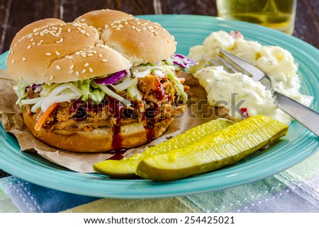 Pulled pork barbeque sandwich with coleslaw sitting on blue plate with potato salad and dill pickle spears and beer in background
