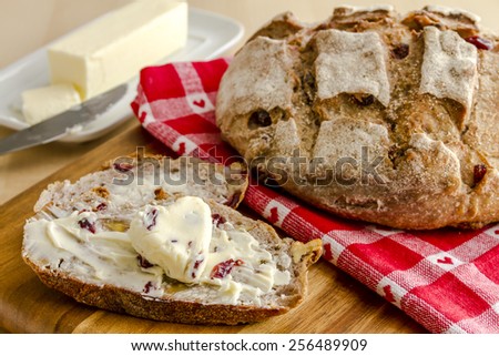 Whole loaf of walnut cranberry bread sitting on red heart napkin on wooden cutting board with slices of bread, butter and knife