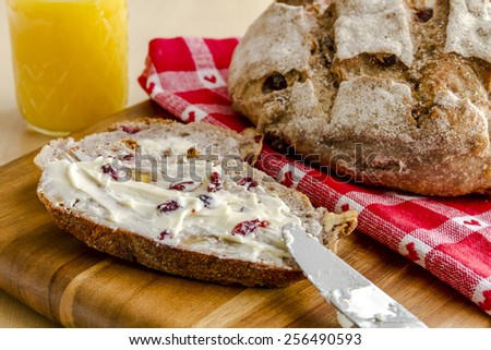 Whole loaf of walnut cranberry bread sitting on red heart napkin on wooden cutting board with slices of bread, butter, knife and glass of orange juice