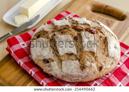 Whole loaf of walnut cranberry bread sitting on red heart napkin on wooden cutting board with butter and knife
