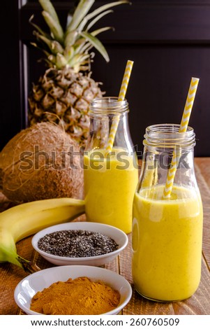 Fresh blended fruit smoothies made with pineapple, banana, coconut, turmeric and chia seeds surrounded by raw ingredients in glass milk bottles with yellow straws