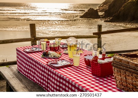 Close up of picnic at the beach overlooking the ocean with haystack rocks at sunset with table set with food, dishes, glasses and red checkered table cloth