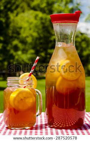 Mason jar mug and tall pitcher filled with fresh brewed iced tea and lemon slices with red swirl straws sitting on a red gingham checked tablecloth on picnic table