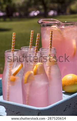Close up of 4 small glass bottles and pitcher filled with fresh squeezed pink lemonade with yellow swirled straws and lemon slices sitting on weather blue drink tray