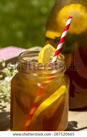 Close up of mason jar mug and tall pitcher filled with fresh brewed iced tea and lemon slices with red swirl straws sitting on a red gingham checked tablecloth on picnic table