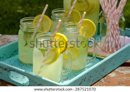 Close up of fresh squeezed lemonade in mason jar mugs and glass pitcher sitting on blue weathered drink tray with pink straws and fresh lemon slices