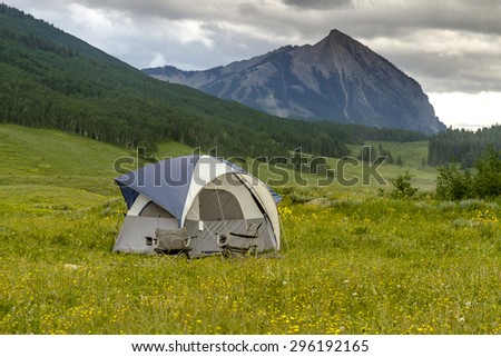 Small camping tent with chairs setup in mountain meadow filled with wildflowers with high mountain peak in distance