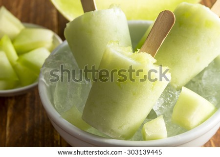 Homemade fresh pureed frozen honey dew melon popsicles in white bowl with ice sitting on wooden table with fresh melon