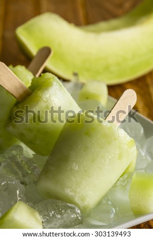 Homemade fresh pureed frozen honey dew melon popsicles on white plate with ice sitting on wooden table with fresh melon pieces