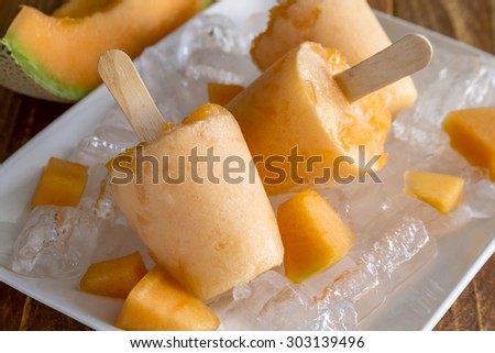 Homemade fresh pureed frozen cantaloupe melon popsicles on white plate with ice sitting on wooden table with fresh melon