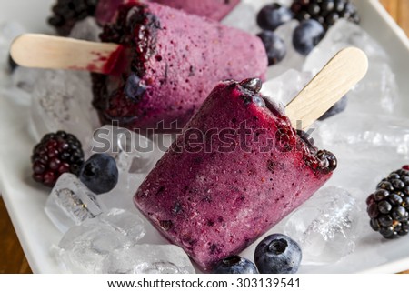 Homemade fresh pureed frozen blueberry and blackberry popsicles on white plate with ice sitting on wooden table with fresh berries