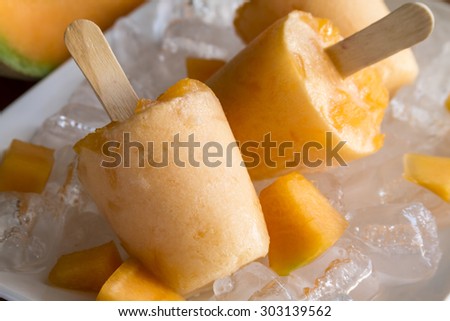 Close up of homemade fresh pureed frozen cantaloupe melon popsicles on white plate with ice sitting on wooden table with fresh melon