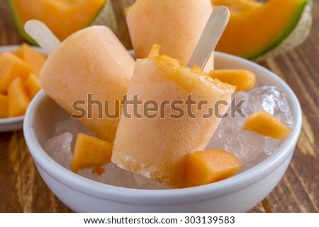 Homemade fresh pureed frozen cantaloupe melon popsicles in white bowl with ice sitting on wooden table with fresh melon
