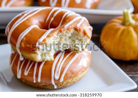 Close up of stack of homemade baked pumpkin donuts with orange pumpkin glaze with bite in top donut sitting on white square plate with small pumpkin in backgound