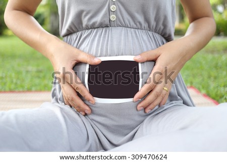 Pregnant women show ultrasound film picture on her belly in the garden.