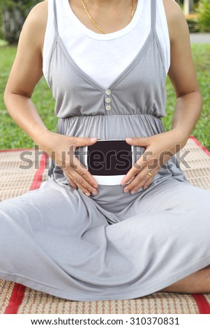 Pregnant women show ultrasound film picture on her belly in the garden.