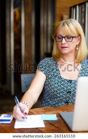 Old senior rich business woman in the glasses sitting at a cafe city with a laptop and calculator, writes notes