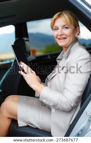 Old senior business woman 60-65 years, sitting in car, holding her Tablet PC computer and looking to camera
