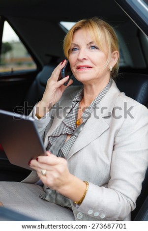 Portrait of happy old senior business woman 60-65 years, sitting in car, holding her Tablet PC computer and talking by mobile phone