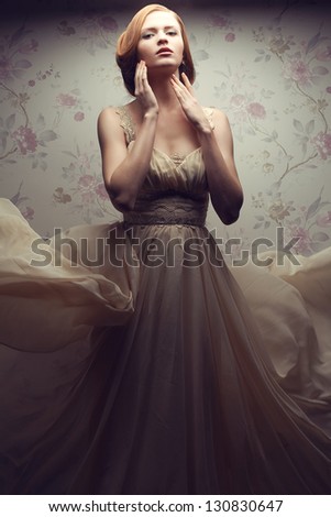 Vintage portrait of a happy glamorous red-haired (ginger) girl posing in great flying beige dress and dancing. Studio shot