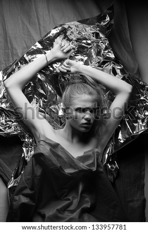 Arty portrait of a fashionable red-haired model with hands up over wrinkled silver foil and gray cloth background. Black and white (monochrome) studio shot