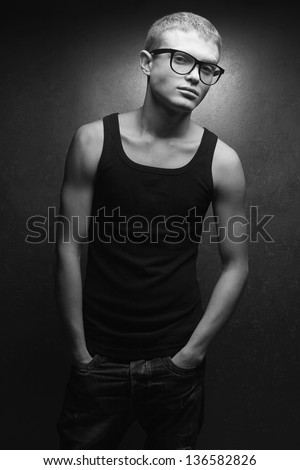 Portrait of a casual young man wearing trendy glasses and posing over gray background with hands in jeans pockets. Hipster style. Black and white (monochrome) studio shot