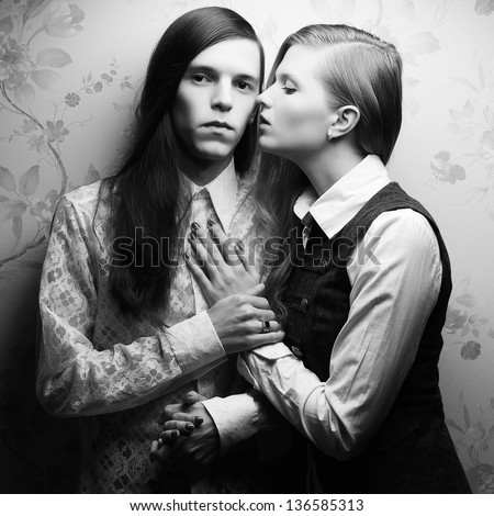 Vintage portrait of beautiful long haired people: handsome boy with brown hair and whispering gorgeous blonde girl posing together. Retro (classic) style. Black and white (monochrome) studio shot.