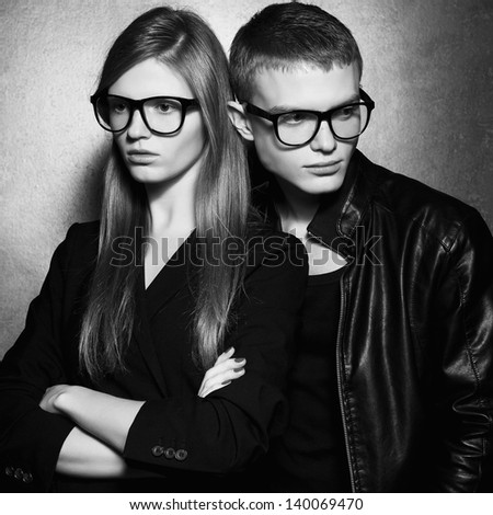 Portrait of gorgeous fashion twins in black clothes wearing trendy glasses and posing over metal background together. Perfect hair and skin. Natural make-up. Hipster style. Black and white studio shot