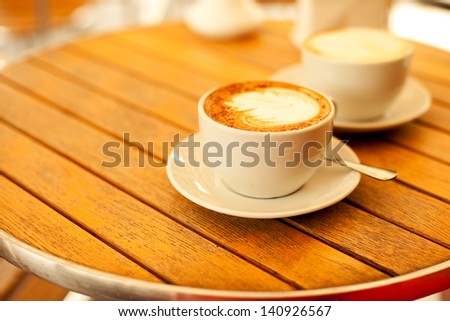Latte art concept. Two cups with cappuccino (hot coffee with milk foam) and canella (cinnamon) on wooden table at street cafe (coffe bar). Vintage style. Copy-space. Outdoor shot.