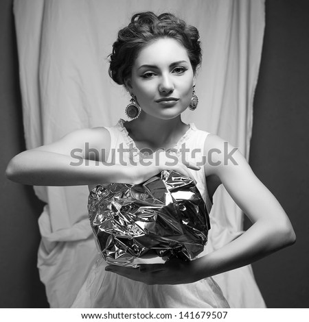 Arty portrait of a fashionable queen-like model holding silver foil sphere over white curtain background. Black and white (monochrome) studio shot