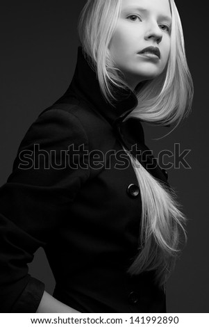 Portrait of a beautiful fashionable model with natural blond hair and great make-up in trendy black coat posing over dark gray background. Black and white (monochrome) studio shot