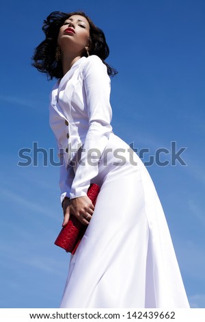 Portrait of a beautiful woman (fashion model) posing in elegant white atlas cocktail dress with red leather clutch in her hands over blue sky background. Sunny & cloudy weather. Outdoor shot