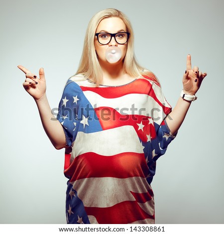 American Dream (Lifestyle) Concept: Young pregnant woman in american flag like dress and trendy glasses chewing bubble gum and dancing over gray background. Hipster style. Studio shot
