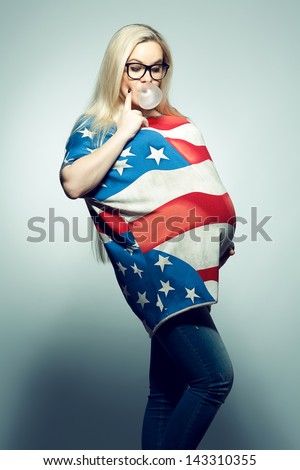American Mom Concept: Young pregnant woman in american flag like dress and trendy glasses chewing bubble gum and looking at her belly over gray background. Hipster style. Studio shot