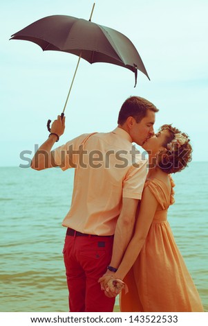 Summer vacation concept. Couple standing on beach near water, holding black umbrella and kissing each other. Hipster style. Outdoor shot