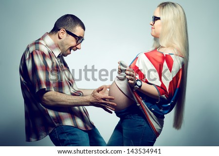 Happy pregnancy concept: portrait of two funny hipsters (husband and wife) in trendy glasses and clothes. Wife showing her belly and husband screaming. Studio shot