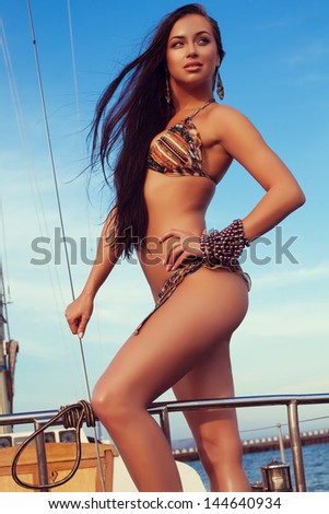 Portrait of a gorgeous long-haired brunette in stylish swimsuit with awesome tan standing on the aft of a sailing yacht. Sunny summer day. Bikini fashion. Outdoor shot
