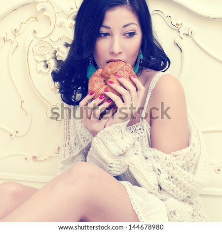 Guilty pleasure concept. Portrait of a young beautiful woman eating her french croissant and thinking something about in a bedroom. Vintage (classic) style. Indoor shot