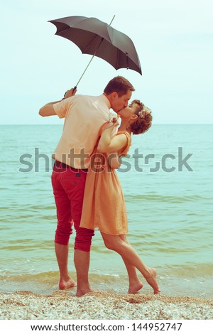 Summer vacation concept. Couple standing on beach near water, holding black umbrella and kissing each other. Hipster style. Happy together. Outdoor shot
