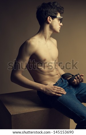 Beautiful (handsome) muscular male model with nice abs in jeans posing in trendy glasses. Boy sitting on a wooden cube. Hipster style. Copy-space. Fashion studio portrait