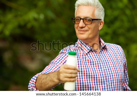 Healthy food concept. Portrait of a mature (old) man in trendy casual shirt and glasses posing in the park and holding a bottle with milk. Sunny summer day. Copy-space. Outdoor shot