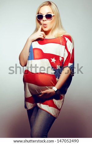 American Mom Concept: Surprised young pregnant woman in american flag like dress and trendy sunglasses posing over gray background. Hipster style. Studio shot