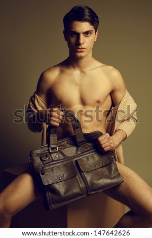 Male high fashion concept. Portrait of a handsome male model sitting on a wooden cube in trendy cardigan, holding leather bag. Perfect skin & haircut. Vogue style. Studio shot