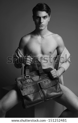 Male high fashion concept. Portrait of a handsome male model sitting on a wooden cube in trendy cardigan, holding leather bag. Perfect skin & haircut. Vogue style. Studio shot