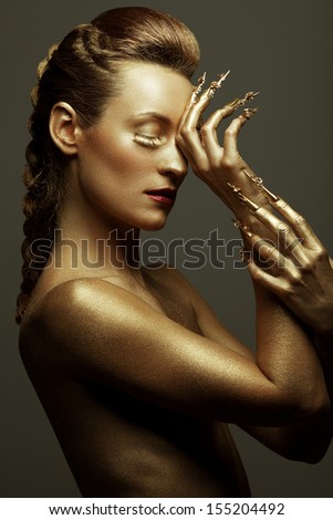 Golden statue of Valkyrie concept. Arty portrait of model with golden healthy skin and shiny lashes. Perfect sporty body. Golden long nails. Profile. Studio shot