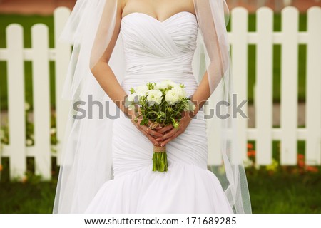 Bride\'s hands with wedding bouquet of beige flowers over white wooden fence and green lawn. Trendy dress with vapory veil. Country (vintage) style. Close up. Copy-space. Outdoor shot